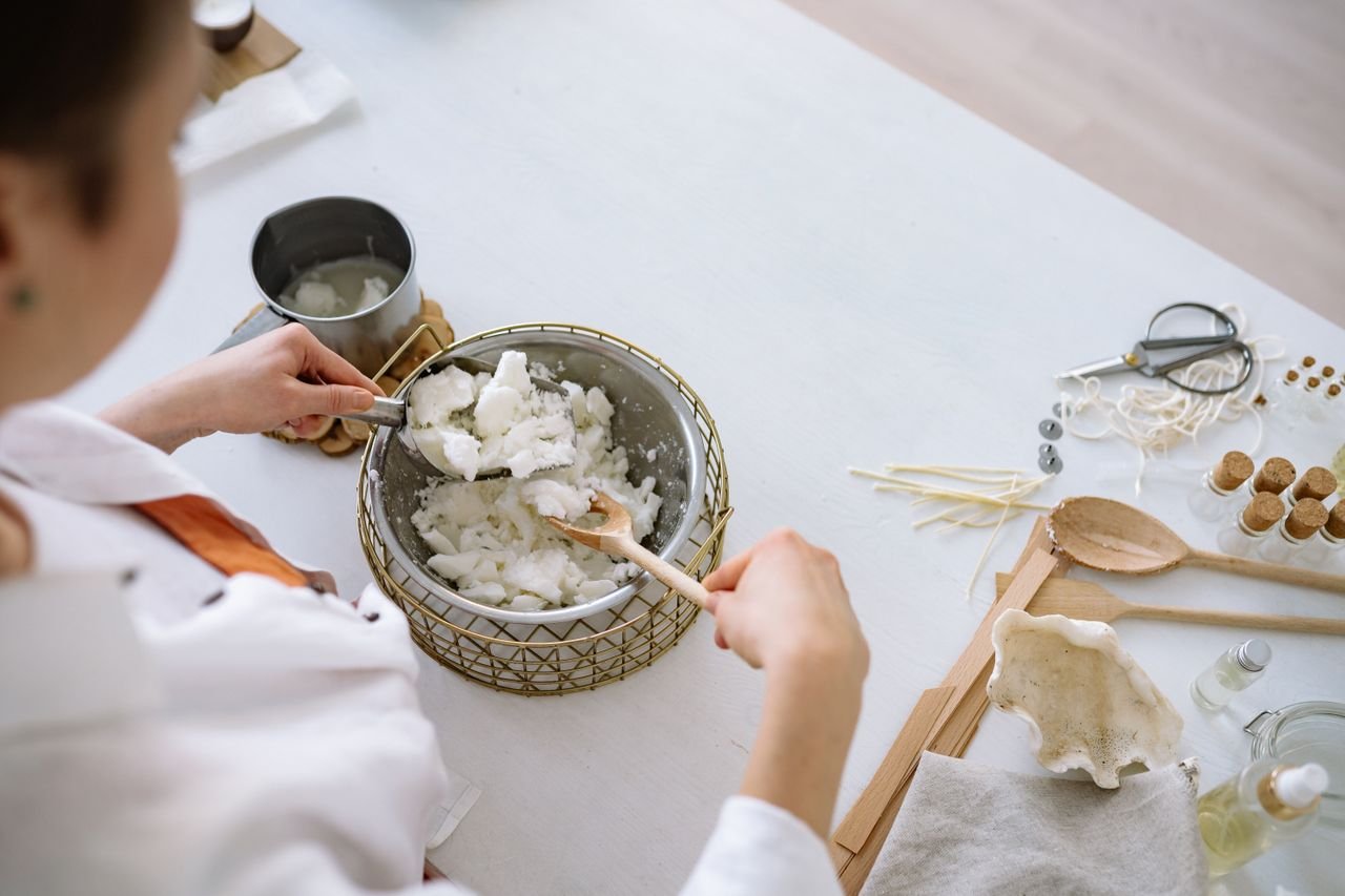 The Alchemy Person melting wax for candle making with tools on a white workspace.